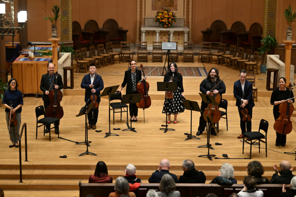 8 Criers stand on a brightly lit stage holding their instruments while audience applauds
